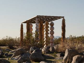 An outdoor art installation in a desert with rocky terrain, consisting of a wooden pergola from which large spheres and conical shapes hang vertically, forming symmetrical chains. Dry plants hang in the corners of the structure, while bushes and mountains are in the background.